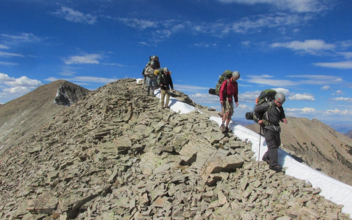 A group of people carrying backpacks and wearing safety gear hike along a rocky ridge with blue skies above.